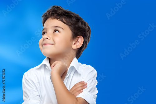 Cheerful Greek child deep in thought, gently touching chin, contemplating against vibrant blue and white background.