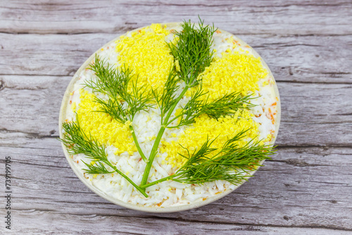 Traditional russian layered salad Mimosa decorated with sprigs of mimosa (made of eggs yolk and dill) on wooden table