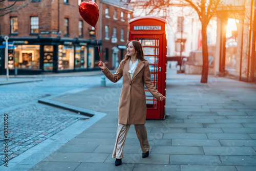 young happy woman with a heart-shaped balloon falling a love, having a fun day, walking against the red phone box in English city Spring is in the air Lifestyle, tourism, valentines day concept