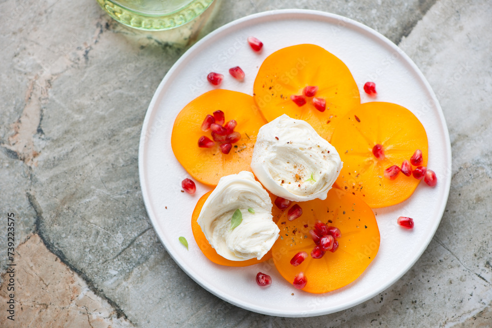 Persimmon slices with mozzarella and pomegranate seeds on a white plate, horizontal shot on a grey granite background, high angle view Persimmon slices with mozzarella and pomegranate seeds on a white plate, horizontal shot on a grey granite background, high angle view