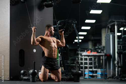 Strong sportsman kneeling, while doing exercise for biceps in gym. Full body of muscular male athlete wearing black shorts, pulling cables of training apparatus indoors. Concept of sport, workout.