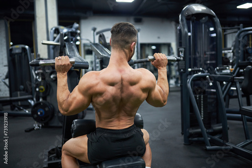 Muscular male bodybuilder exercising with training apparatus in gym. Back view of anonymous strong shirtless man, building back muscles, working out at evening. Sport, lifestyle, bodybuilding concept.