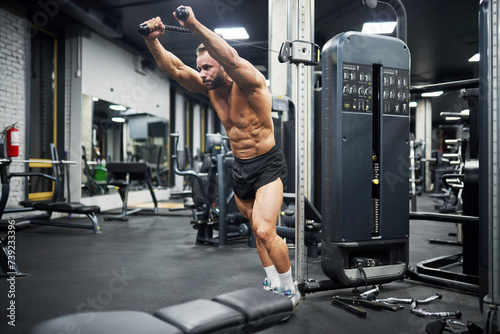 Middle-aged Caucasian man training in crossover machine indoors. Side view of bearded crossfit male athlete doing pull-downs, lifting metal plates of cable crossover machine in gym. Crossfit concept.