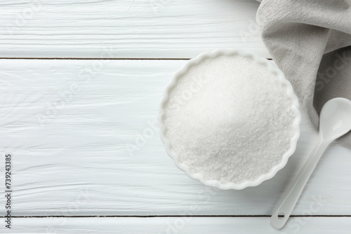 Granulated sugar in bowl and spoon on white wooden table, top view. Space for text