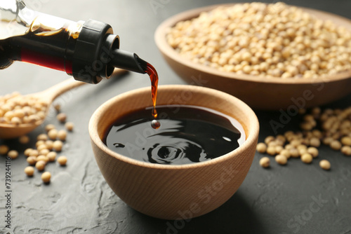 Pouring tasty soy sauce from bottle into bowl on black table, closeup photo
