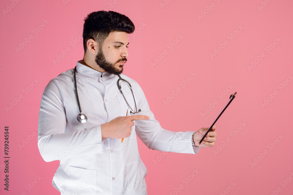 Side view of brunette doctor, intern holding folder, looking, pointing by finger, posing indoors. Isolated on pink studio background. Side view of brunette doctor, intern holding folder, looking, pointing by finger, posing indoors. Isolated on pink studio background.