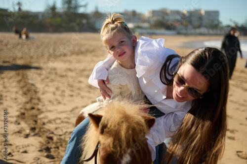 Smiling mother stroking a little pony while supports her daughter on horse riding. People. Active healthy lifestyle.