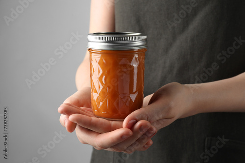 Woman holding glass jar of delicious persimmon jam on gray background, closeup. Space for text photo