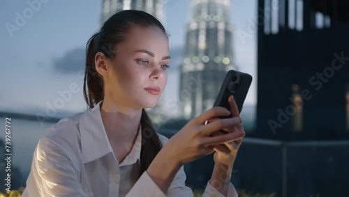 A woman looks at her phone, smiles and types a message against a backdrop of a night city and glowing buildings, work and leisure.