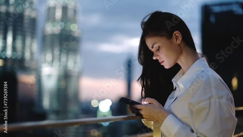 A woman looks at her phone, smiling and laughing typing a message backdrop of a nighttime city and glowing buildings on the roof of a skyscraper, hair flying in the breeze, work and leisure city life.