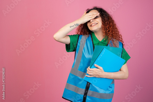 Front view of pertty, female paramedic wearing vest, standing, raising hand, closing eyes, smiling. Isolated on pink studio background. photo