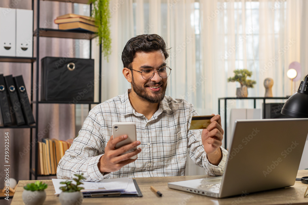 Happy Hispanic businessman using bank credit card and smartphone for online shopping payments at home office. Business transaction. Smiling guy freelancer purchase with cellphone E-banking app service