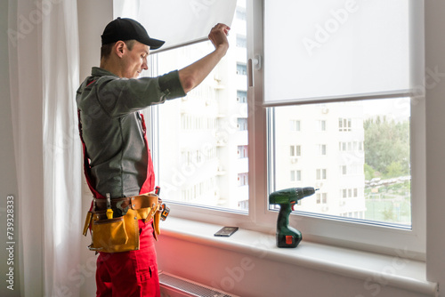 Maintenance man is fixing windows in a living room using a skrewdriver. He is wearing glasses, yellow gloves and a black apron photo