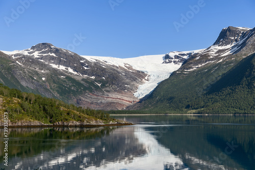 A serene fjord cradles the reflection of the majestic Svartisen Glacier, flanked by lush, green slopes under the bright Nordic sky