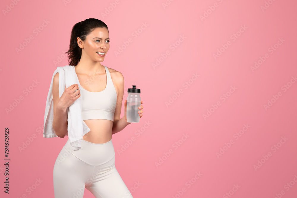 A smiling fitness enthusiast in white attire carries a water bottle and a towel over her shoulder A smiling fitness enthusiast in white attire carries a water bottle and a towel over her shoulder