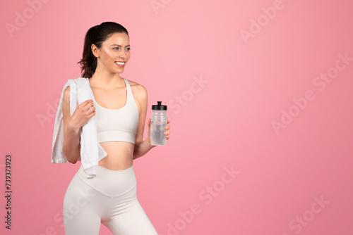 A smiling fitness enthusiast in white attire carries a water bottle and a towel over her shoulder photo