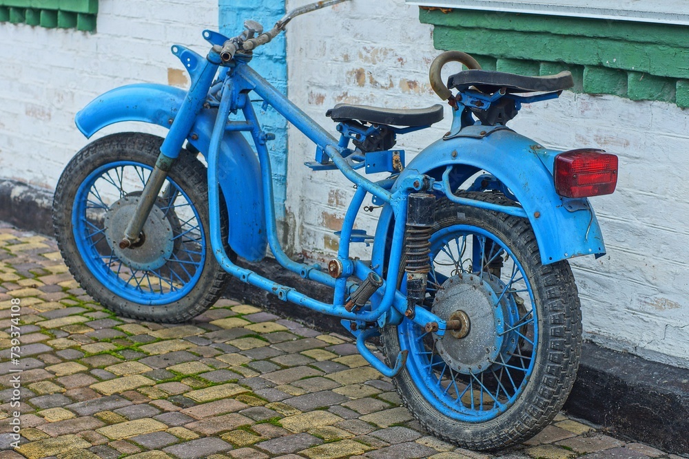 an old blue iron frame with a saddle with wheels and wings and a steering wheel from a retro heavy motorcycle stands on a concrete floor near a white wall during the day on the street