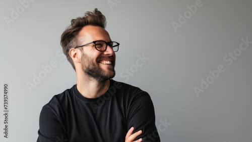 A young man with a beard and glasses is wearing a black t-shirt, smiling and looking away from the camera. The background is a light gray color