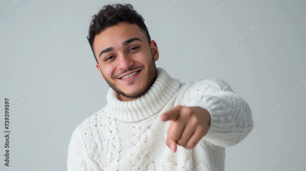 Confident young man with dark hair and a well-groomed beard, wearing a white turtleneck sweater, smiling and pointing at the camera on grey background - obrazy, fototapety, plakaty 