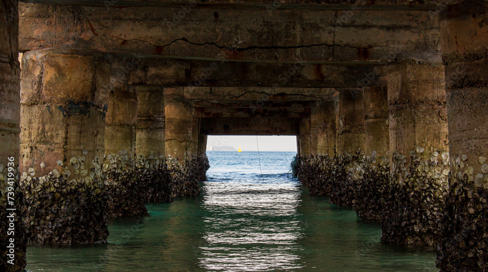 Perspective of the blue water from the coast beneath the concrete pier. View of the turquoise sea at the coast from beneath the concrete pier.