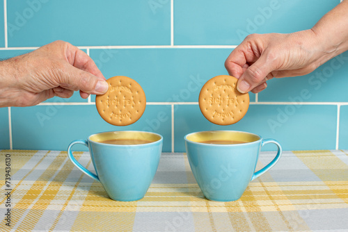 Cups of tea in attractive blue and yellow cups with hands dunking rich tea biscuits. Cups isolated against a blue tiled background and sitting on yellow check. Mugs of hot drink with a sweet treat. 
