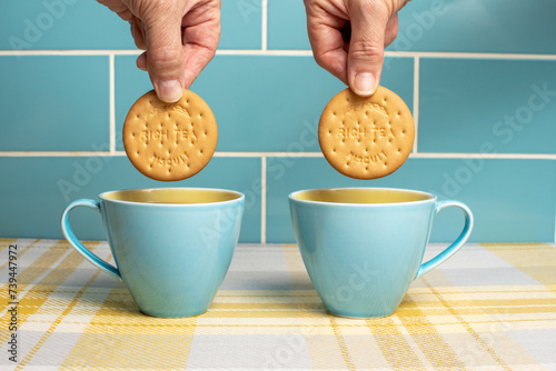 Obraz na płótnie Cups of tea in attractive blue and yellow cups with hands dunking rich tea biscuits