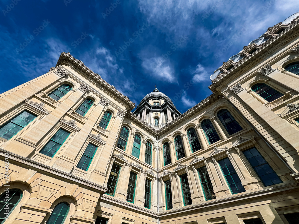 Fototapeta premium Rear views of the Illinois State Capitol Building in Springfield, Illinois, USA. Cloudy blue skies overhead. The US flag, State of Illinois Flag, and Black History Month Flag fly atop the dome.