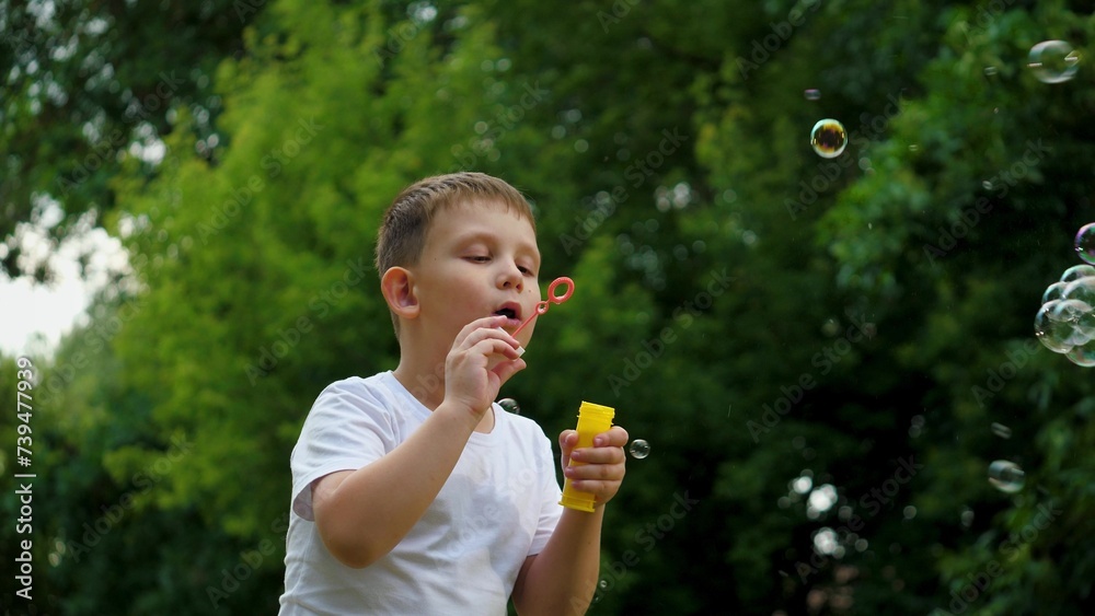 Little boy blows soap bubbles walking in green city park. Happy little