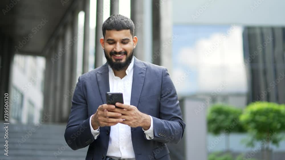 A smiling businessman is using a phone while walking along the street near an office building. Happy handsome bearded male texting, chatting online, browsing social media web, reads writes messages A smiling businessman is using a phone while walking along the street near an office building. Happy handsome bearded male texting, chatting online, browsing social media web, reads writes messages