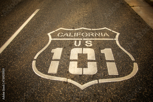 Highway 101 sign painted on the black asphalt road with city diffused in the background. The shot is from Oceanside California