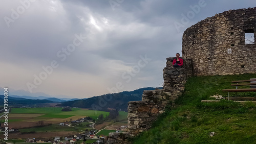 Woman sitting on medieval stone walls. Scenic view of castle ruins Burgruine Griffen in Voelkermarkt, Carinthia, Austria. Tourist attraction on cloudy day. Historic travel destination photo