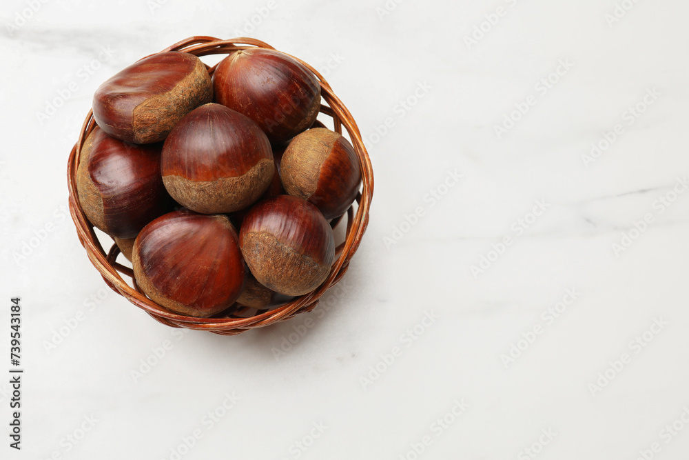 Obraz premium Sweet fresh edible chestnuts in wicker bowl on white marble table, top view. Space for text