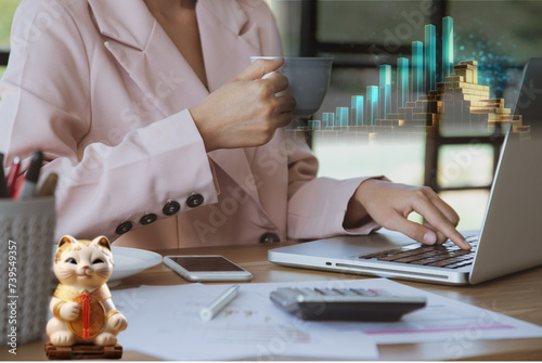 Mujer de negocios trabajando en su laptop tomando su taza de café, sobre el escritorio vemos su móvil, su calculador y su gato de la suerte. Un gráfico de barras muestras sus ganancias. photo