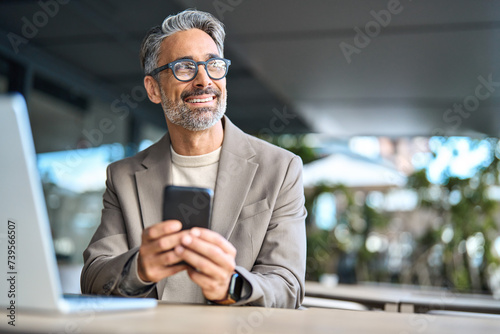 Happy middle aged business man executive using mobile phone sitting outside office. Stylish older busy businessman investor wearing glasses holding smartphone looking away at copy space with cellphone
