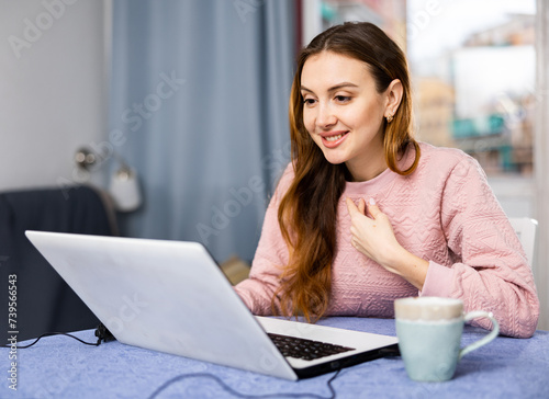 Young positive woman sitting at a table in an apartment communicates with someone via video link on a laptop