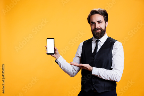 Male waiter shows white screen in studio, presenting isolated copyspace template on smartphone display. Professional elegant butler holding empty mockup screen over yellow background.