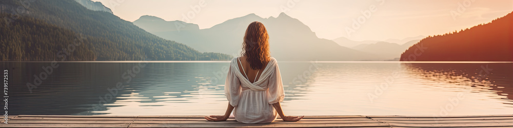 Young woman from rear side meditating by the lake in summer sunnset. Practicing mindfulness and meditation in a peaceful nature.