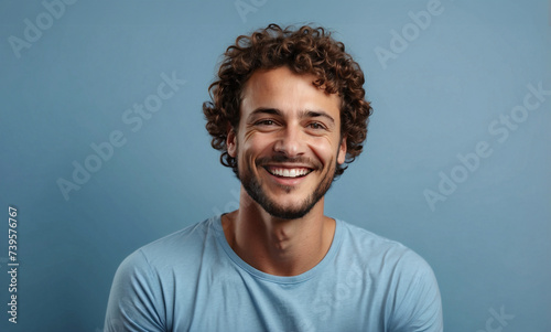 Portrait of handsome 30 year old brown curly haired guy with wide smile and light blue t-shirt, looks at camera with happy expression