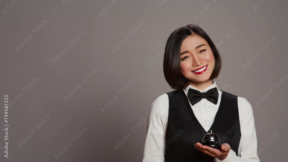 Woman hotel employee ringing front desk service bell in studio, working as a receptionist and registering guests in rooms. Asian concierge presenting a bell to call for assistance. Camera B. Woman hotel employee ringing front desk service bell in studio, working as a receptionist and registering guests in rooms. Asian concierge presenting a bell to call for assistance. Camera B.
