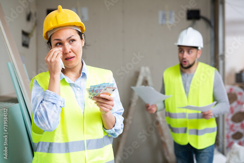 Upset crying adult construction workwoman frustrated with little earnings standing at building site with banknotes in hands.. photo