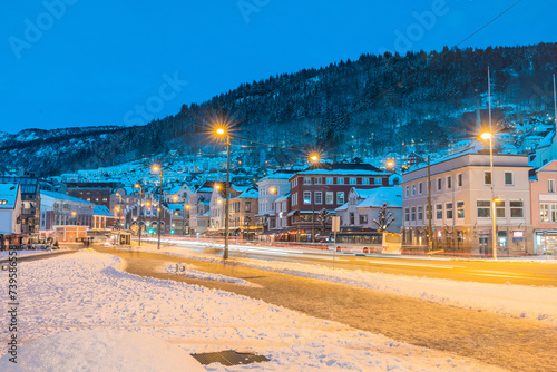 Main street in bergen ar night, road covered in snow and ice. Colorful houses aroud lit by streetlamps and snow capped hill in the background. Lovely city.