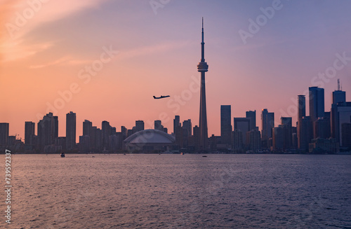 Summer evening view across a stretch of Lake Ontario's Inner Harbour with a passenger airplane taking off against the silhouettes of downtown Toronto skyscrapers at dusk under a hazy sky with pink and