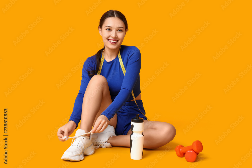 Naklejka premium Young woman in sportswear and with measuring tape tying shoelaces on yellow background. Weight loss concept