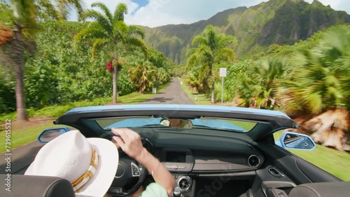 Happy girl in blue convertible car enjoying summer vacation, waiving hand out of the car and showing shaka hawaiian sign. Woman travels by tropical island with tropical exotic mountain landscape Oahu photo