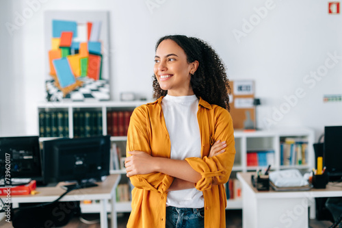 Positive successful confident beautiful hispanic curly haired business woman in orange shirt, business coach, manager, standing in a modern office with arms crossed, looking away and smiling photo