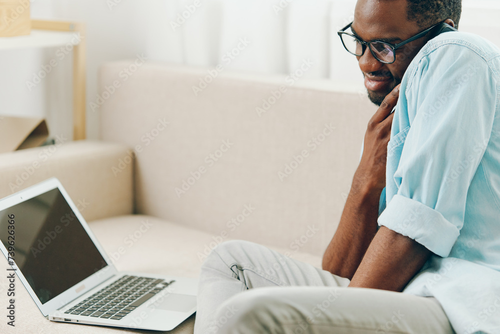 Smiling African American man working on laptop while sitting on a sofa in his modern apartment He is typing on the computer, talking on his phone, and enjoying the convenience of online shopping and