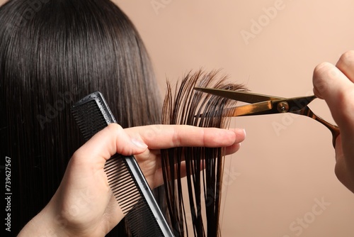 Hairdresser cutting client's hair with scissors on light brown background, closeup