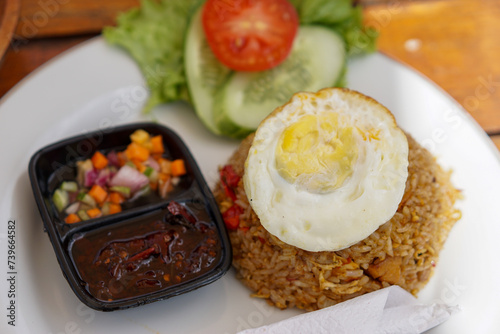 Fried rice served on a white plate topped with egg, spicy sambal and pickles.