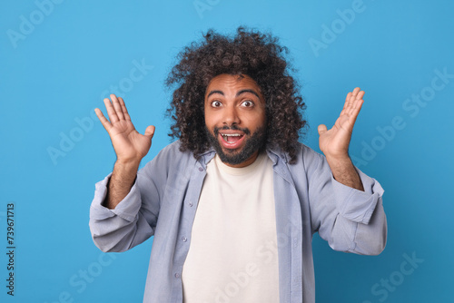 Young shocked cheerful Arabian man with curly hair raises hands in confusion after receiving good news from college friends or work colleagues dressed in casual style stands in blue studio.