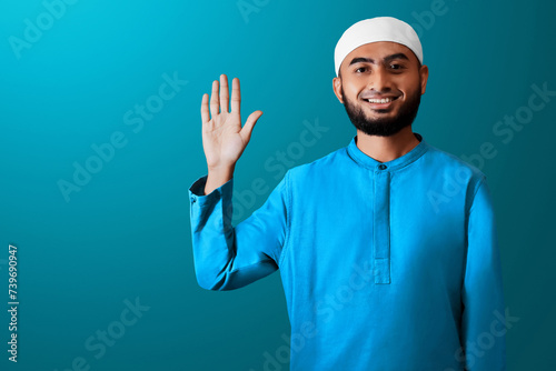 Portrait of handsome young asian muslim man with beard saying hello, waving hand gesture and smiling isolated on blue studio background